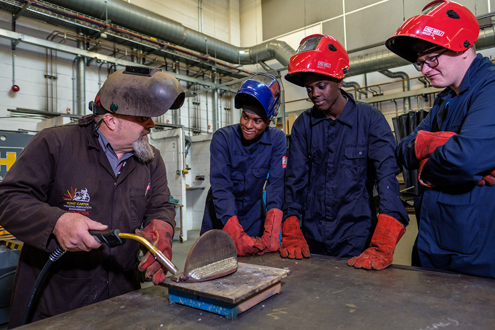 Three young men in overalls watching an older man do welding