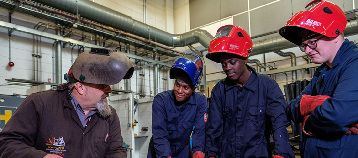 Three teenage boys in overalls watching an older man welding