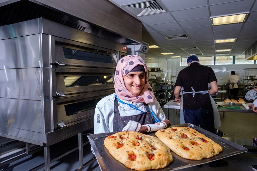 A woman standing in front of big ovens holding a tray with bread on