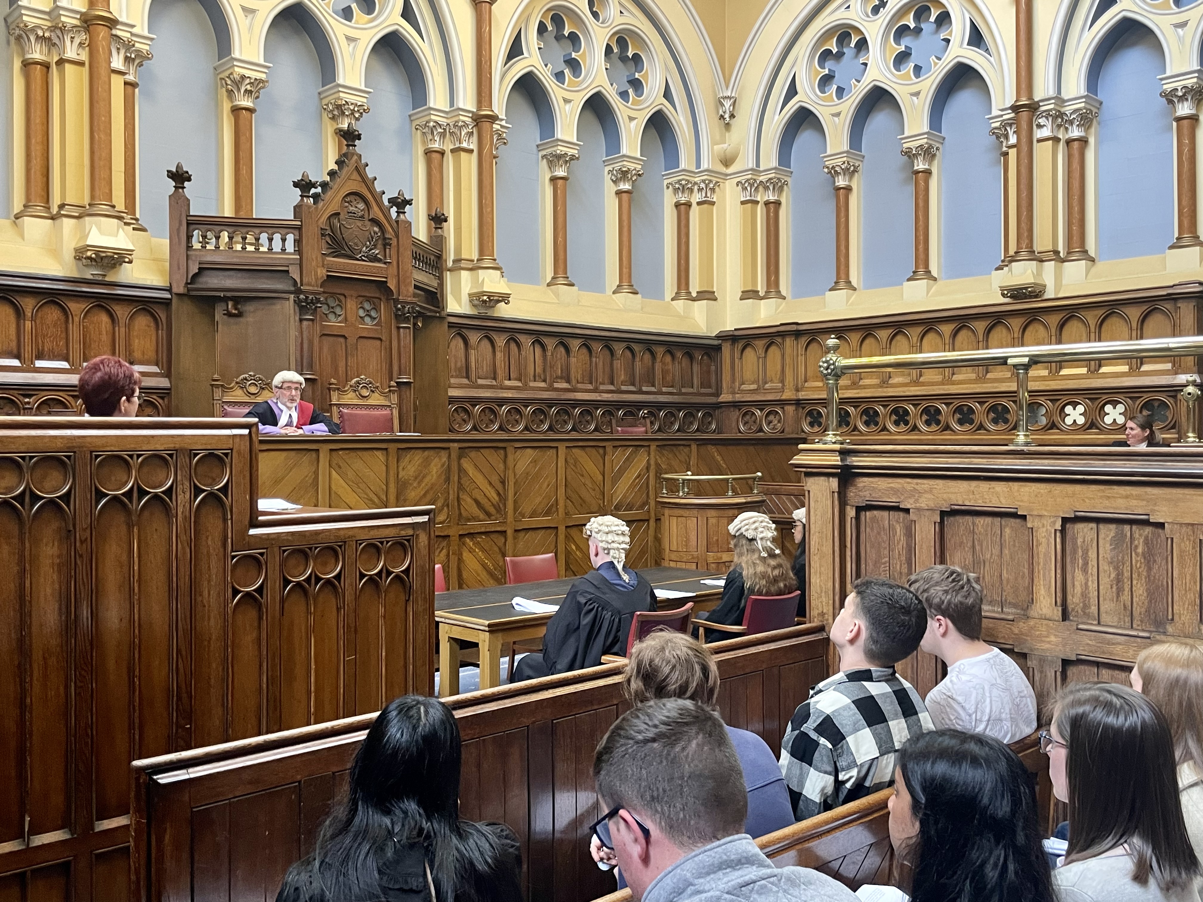 A judge sat at the front of a courtroom, facing two people in barristers' wigs and gowns, with a group of young people watching